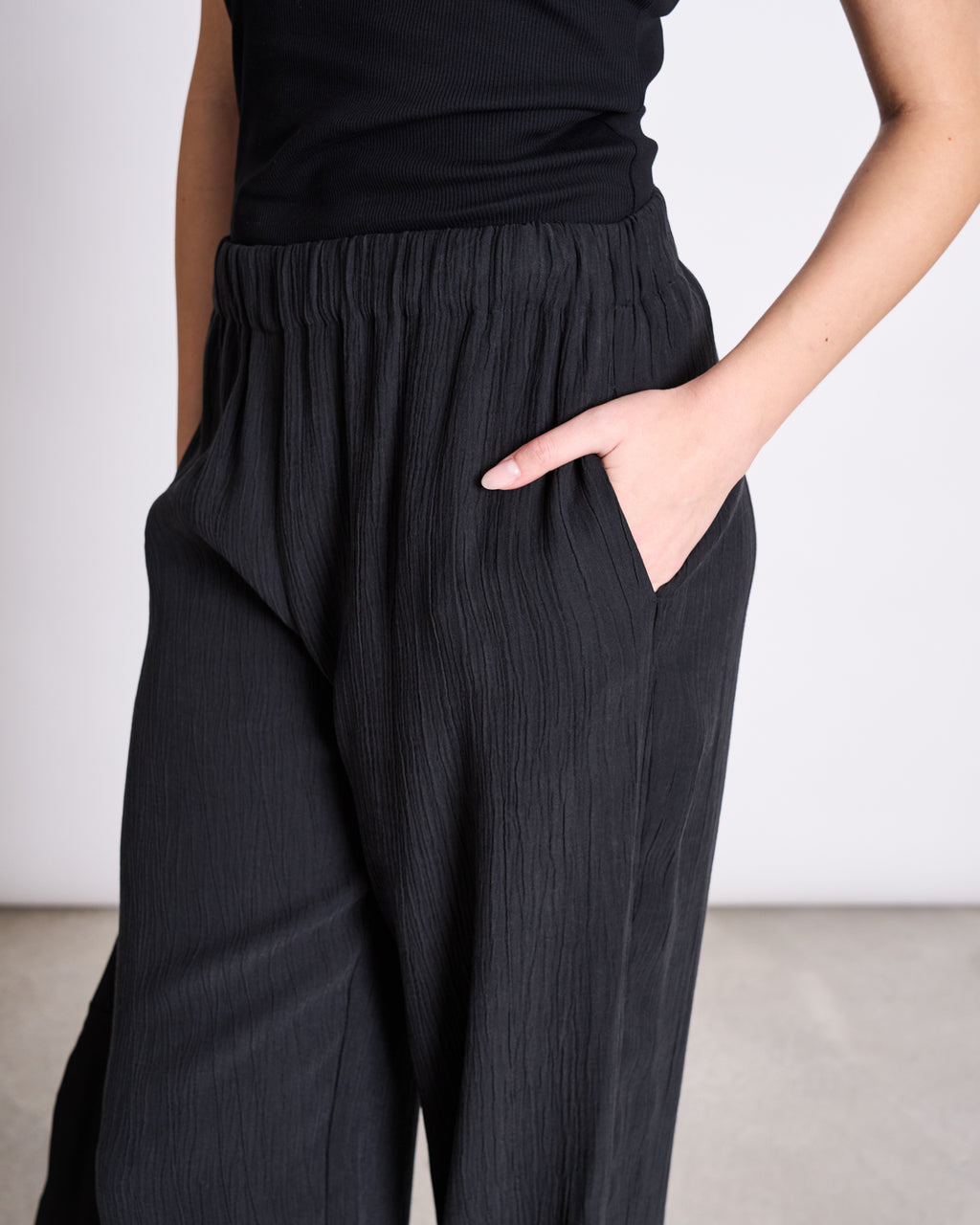A person stands against a plain background wearing jan-n-junes CRINKLE PANTS HAVA BLACK and a black sleeveless top, with one hand in their pocket.