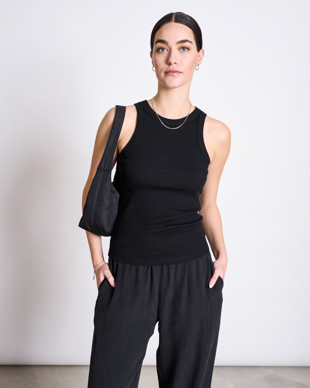 A woman with dark hair pulled back wears the jan-n-june ORGANIC COTTON TANK TOP PORTO LONG BLACK GOTS, paired with loose dark pants and a small black shoulder bag, standing against a plain white background with her hands in her pockets.