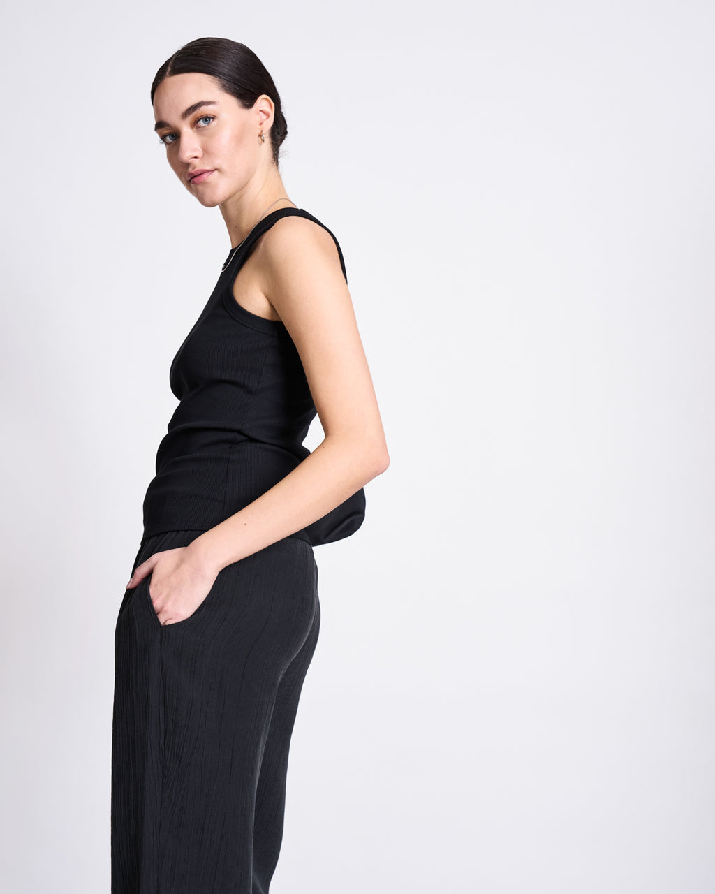 A person with dark hair pulled back wears the jan-n-june ORGANIC COTTON TANK TOP PORTO LONG BLACK GOTS and loose dark pants, standing with one hand in a pocket, looking at the camera against a plain light background.