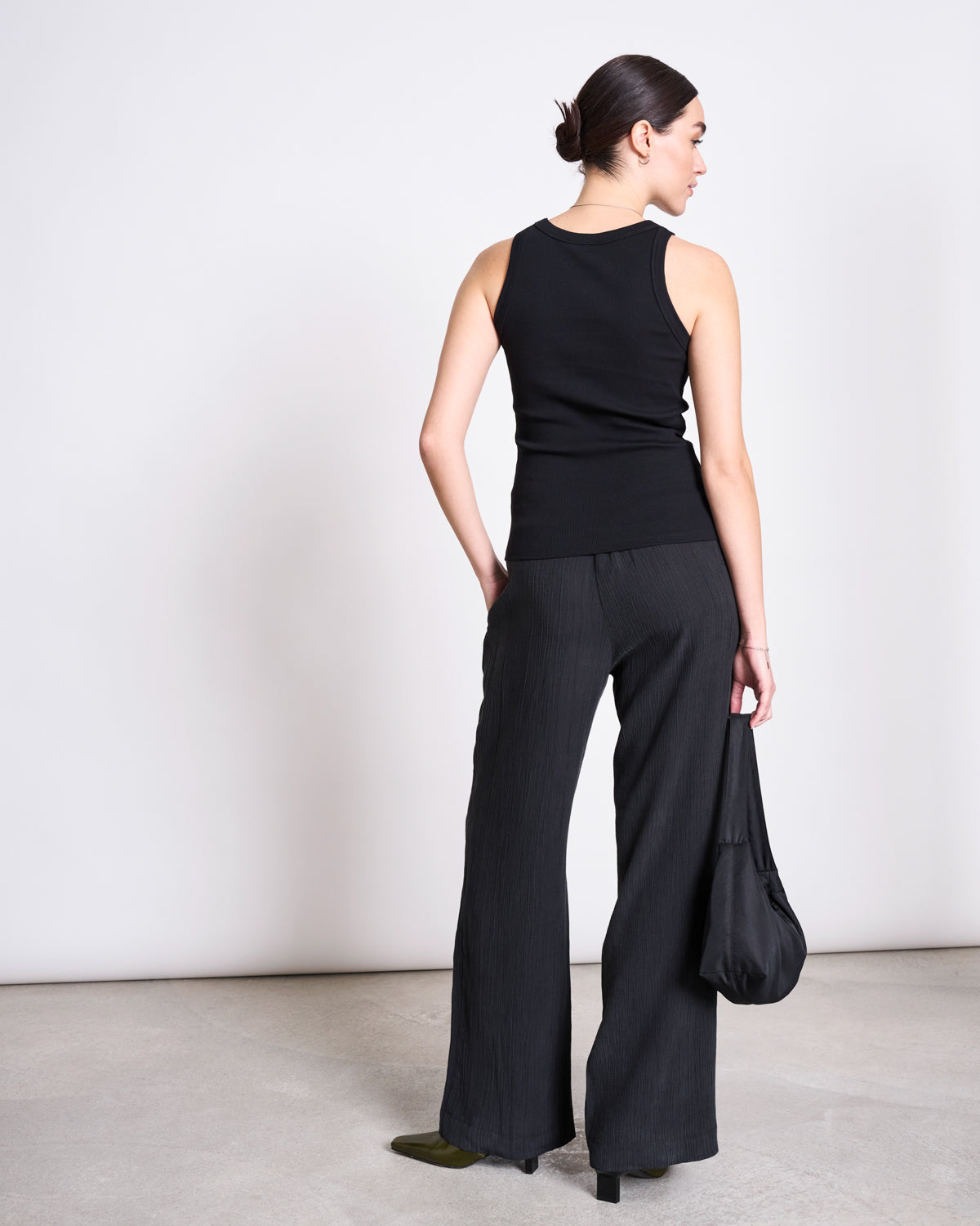 A woman with a neat bun stands on a gray floor against a light wall, wearing the jan-n-june ORGANIC COTTON TANK TOP PORTO LONG BLACK GOTS and loose black pants. She carries a black bag, showcasing stylish everyday comfort.