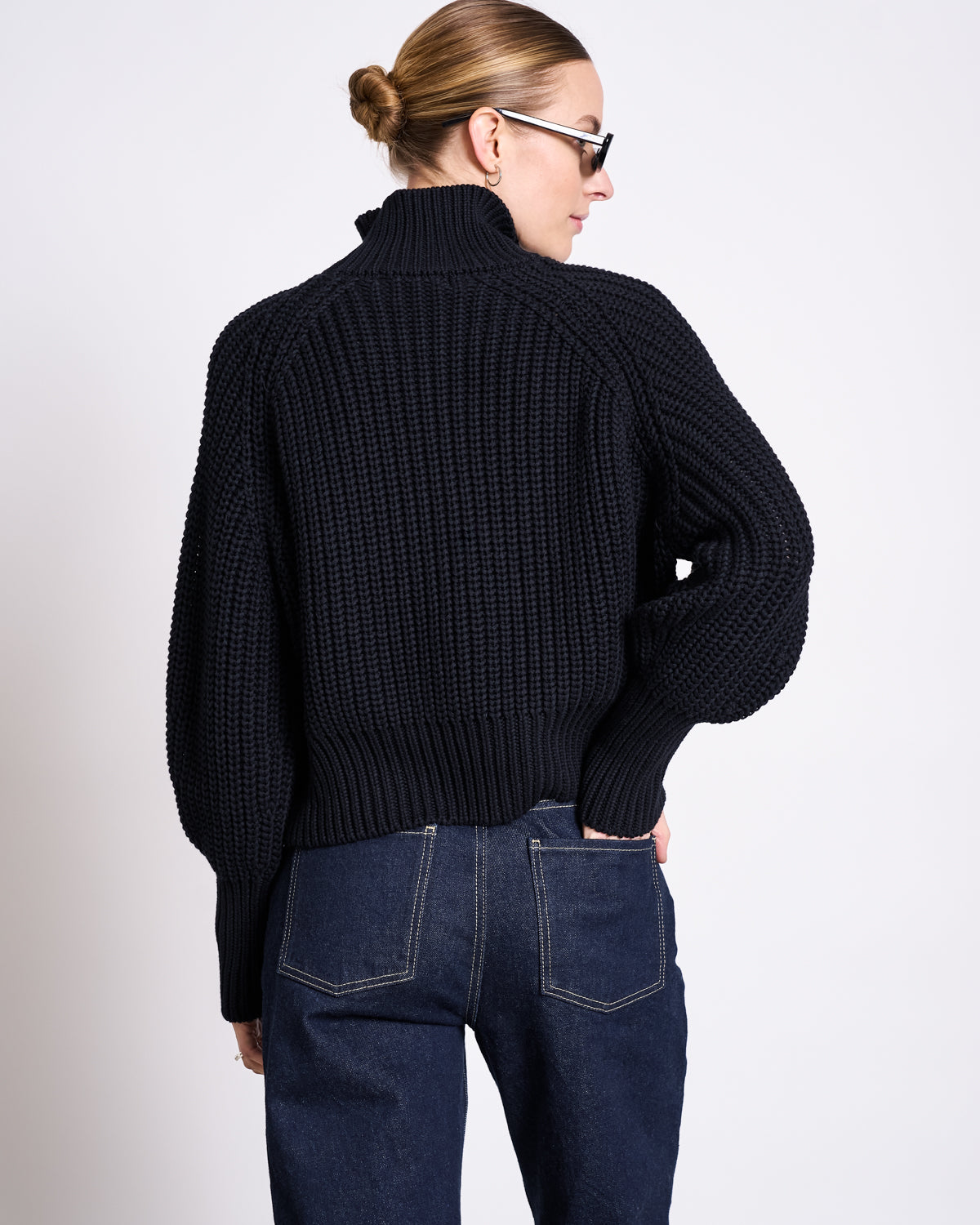 A person with blonde hair in a bun, sunglasses, and a chunky black knit sweater wears jan-n-june’s STRAIGHT LEG HIGHWAIST JEANS VILCA DARK BLUE DENIM GOTS, standing with one hand in their back pocket facing away from the camera.