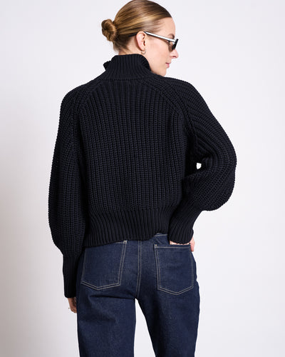 A person with blonde hair in a bun, sunglasses, and a chunky black knit sweater wears jan-n-june’s STRAIGHT LEG HIGHWAIST JEANS VILCA DARK BLUE DENIM GOTS, standing with one hand in their back pocket facing away from the camera.