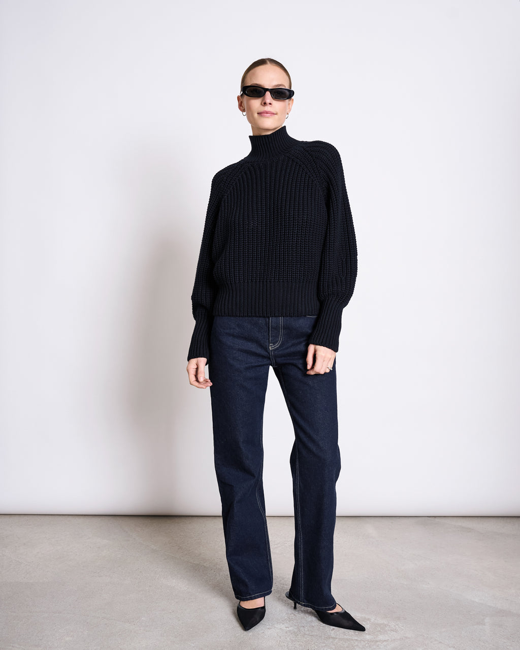 A person stands against a white wall wearing dark sunglasses, a black turtleneck, black pointed-toe shoes, and jan-n-junes STRAIGHT LEG HIGHWAIST JEANS VILCA DARK BLUE DENIM GOTS.