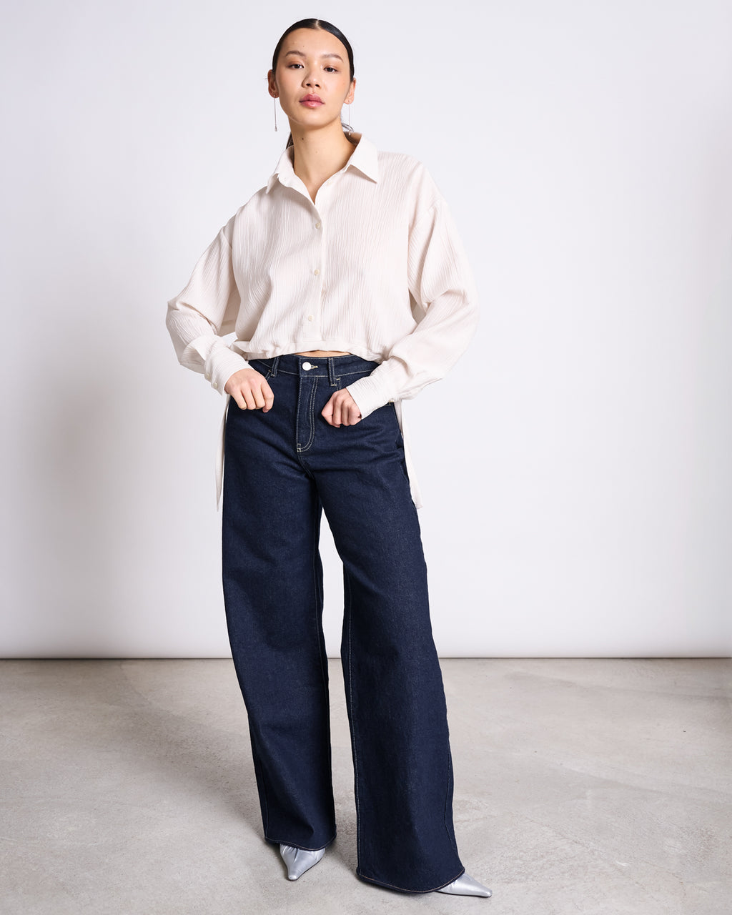 A woman poses against a plain background wearing jan-n-junes WIDE LEG LOWRISE JEANS TACNA DARK BLUE DENIM GOTS, a cream long-sleeve button-up shirt, and silver pointed-toe shoes. She stands facing forward with one hand on her hip.