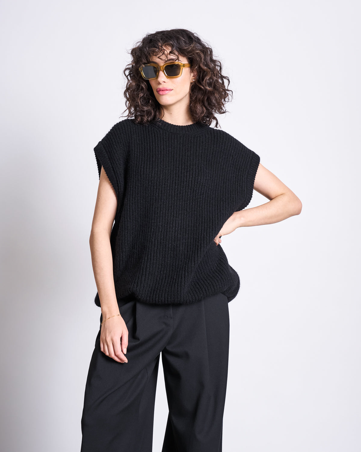 A woman with curly hair and sunglasses poses confidently against a white background, wearing jan-n-junes WOOLY SLIPOVER THANDA BLACK—an oversized Mohair Wool slipover—paired with wide-leg black pants.