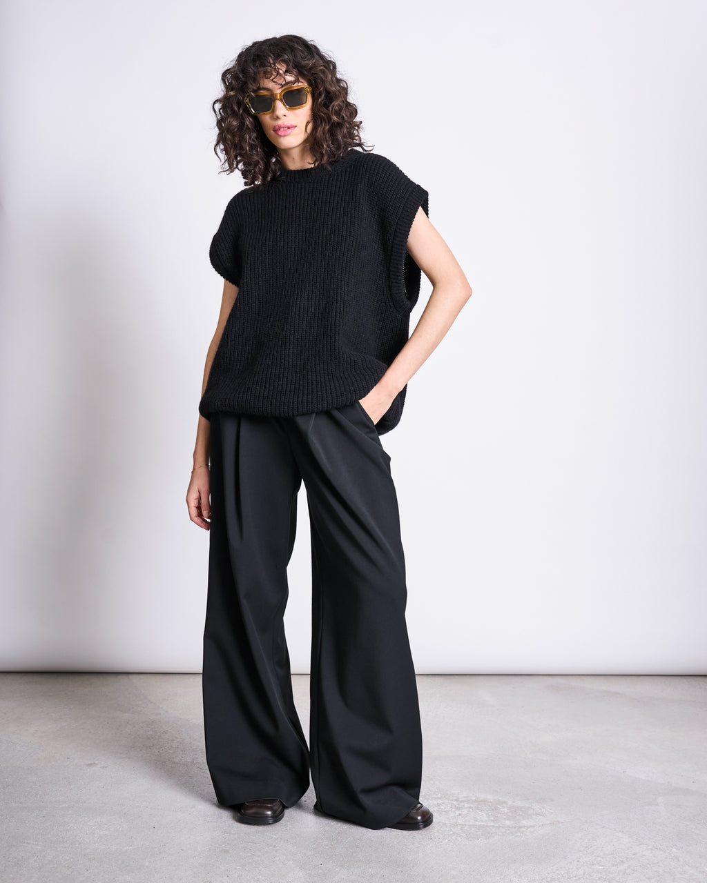 A person with curly hair in sunglasses and a loose black knit top wears jan-n-junes WIDE PANTS GIZA BLACK—high waist, wide leg, recycled polyester—standing against a plain white wall with one hand in their pocket.