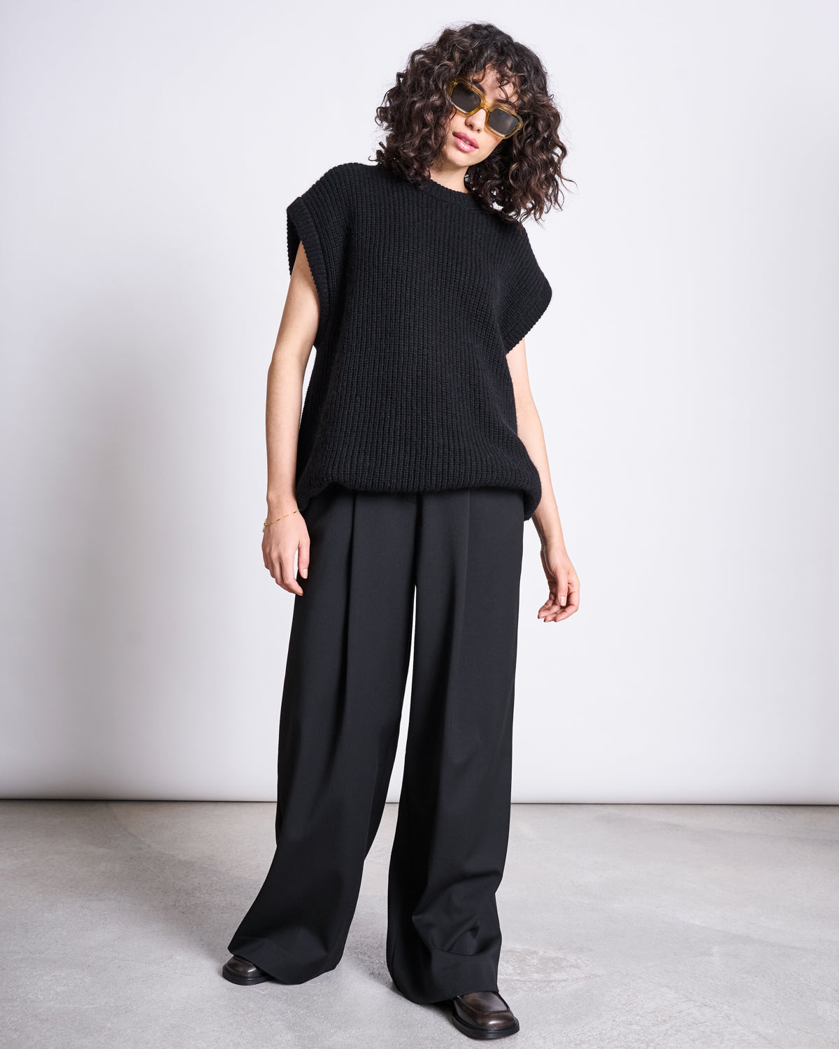 A person with curly hair and sunglasses wears the jan-n-june WOOLY SLIPOVER THANDA BLACK—an oversized black mohair wool slipover—and wide-leg black pants, standing on a light floor against a plain white background.