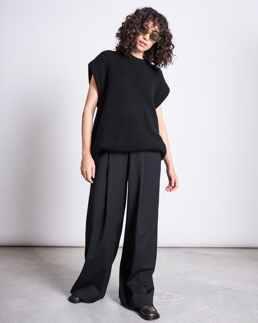 A person with curly hair and sunglasses wears a loose black knit sleeveless sweater and jan-n-june’s WIDE PANTS GIZA BLACK, standing on a light floor against a plain white background.