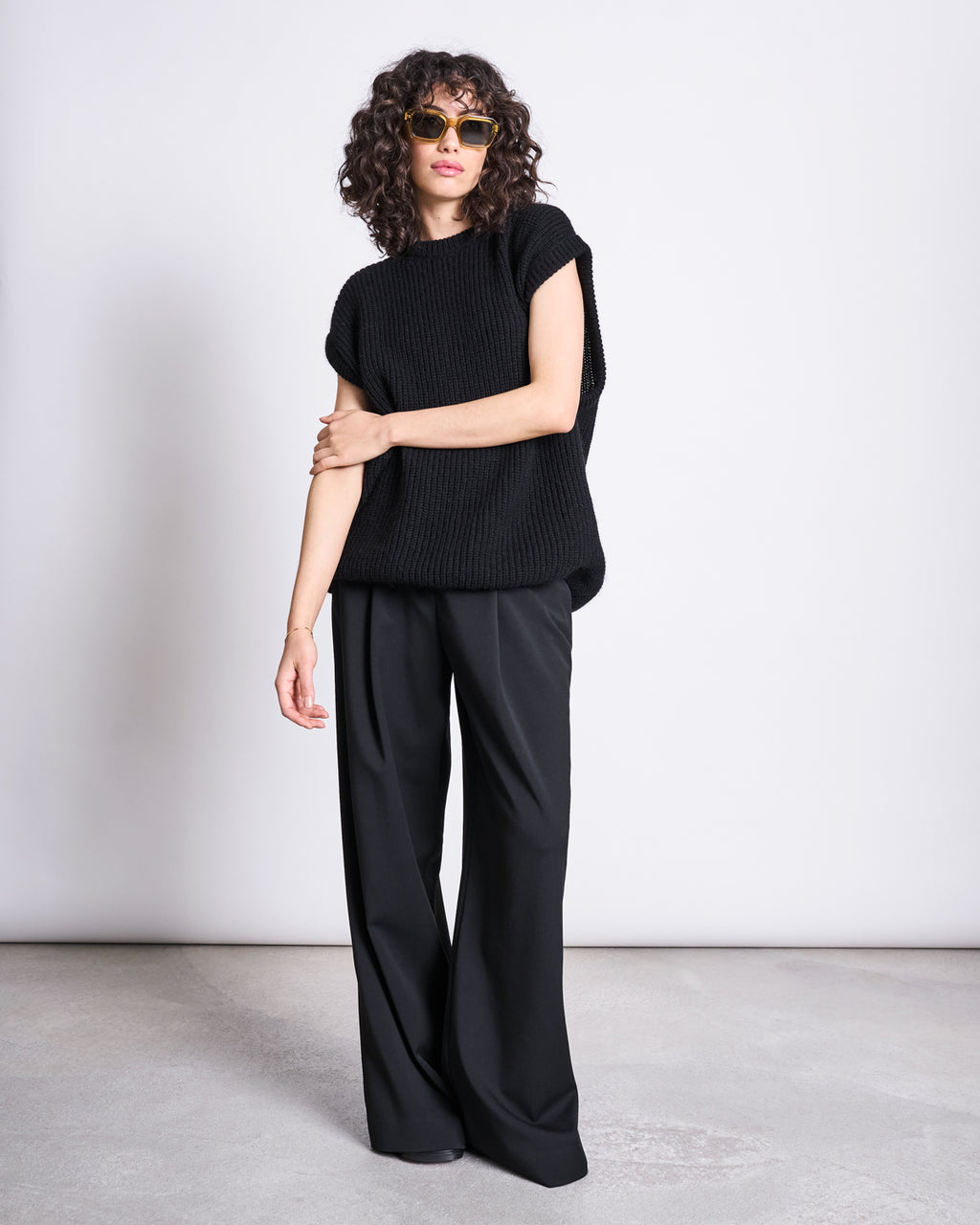 A person with curly hair wears sunglasses and the jan-n-june WOOLY SLIPOVER THANDA BLACK with wide-leg black pants, standing against a plain light background.