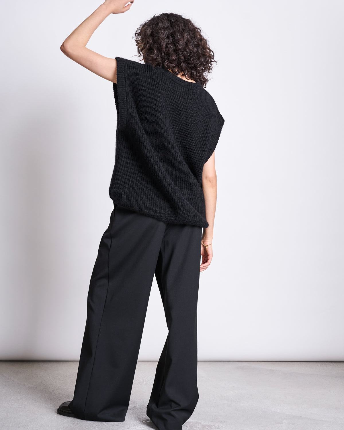 A person with curly hair stands with their back to the camera, wearing an oversized fit jan-n-june WOOLY SLIPOVER THANDA BLACK and wide-leg black pants against a plain light background.