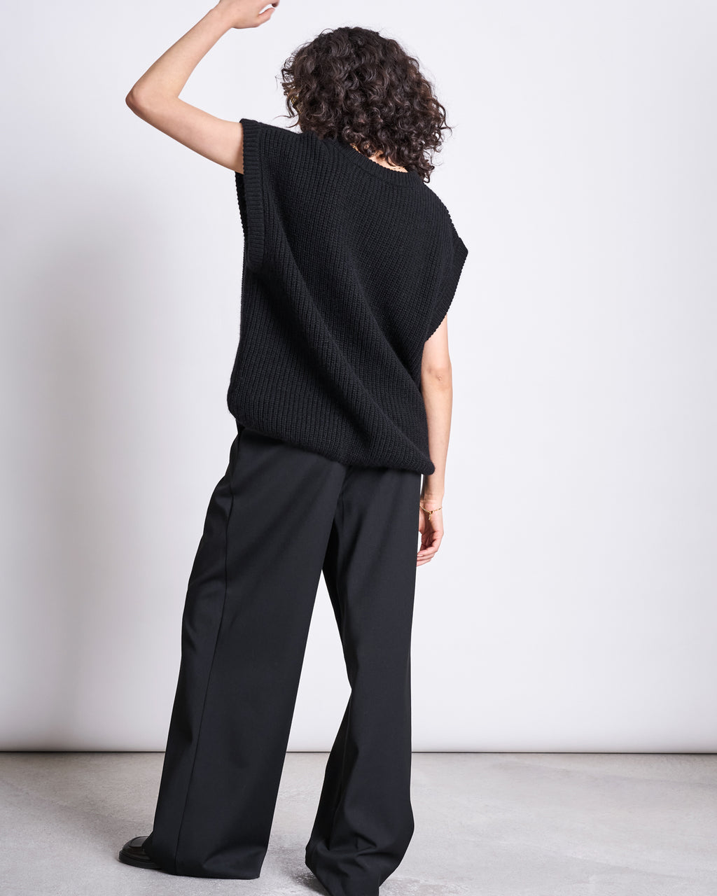 A person with curly hair stands facing away, wearing jan-n-june’s WIDE PANTS GIZA BLACK—high waist, wide-leg black pants made from recycled polyester—paired with a loose black sleeveless sweater. One arm is bent upward against a plain light background.