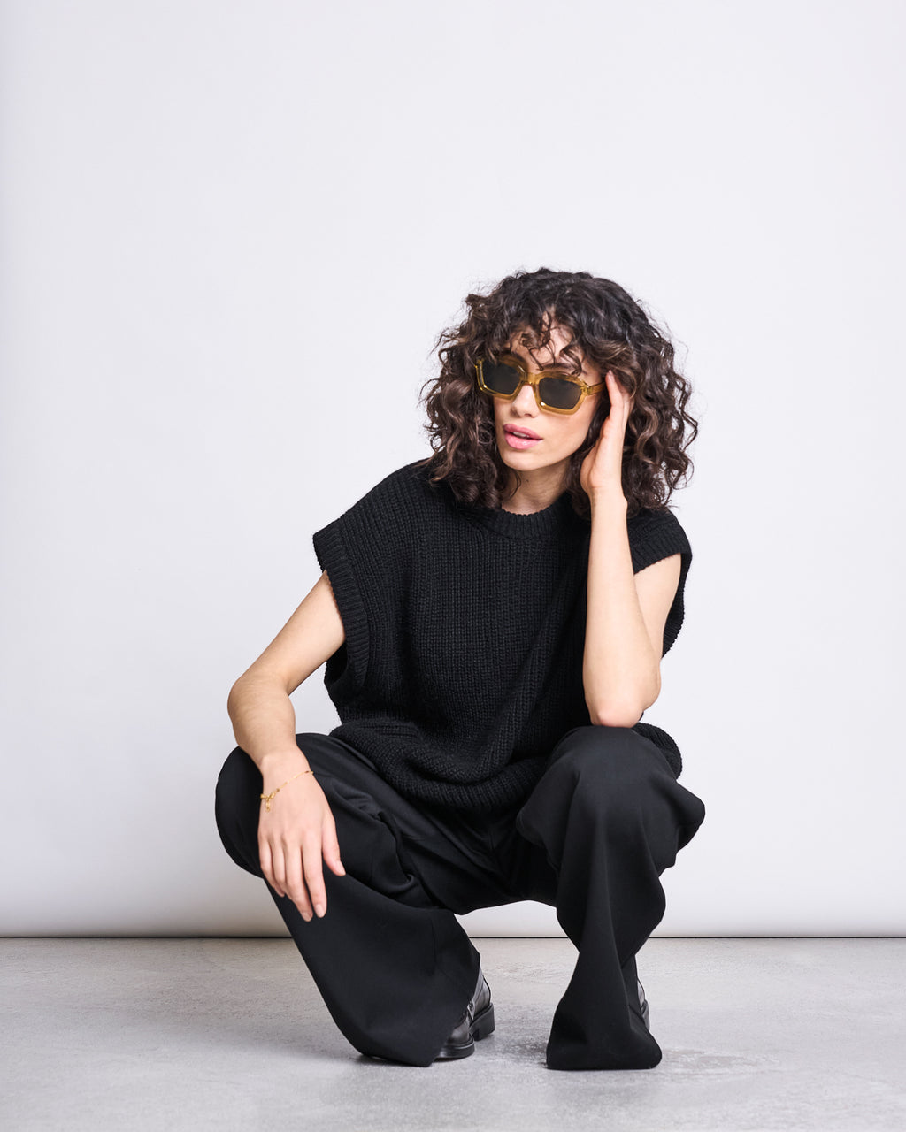A person with curly hair, oversized sunglasses, loose black pants, and the jan-n-june WOOLY SLIPOVER THANDA BLACK in an oversized fit is squatting on the ground against a plain white background.