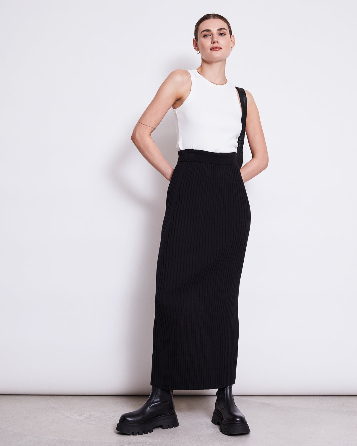 A woman stands against a white wall wearing the jan-n-june MIDI KNIT SKIRT EBBA BLACK GOTS, paired with a sleeveless white top, black chunky boots, and a shoulder strap accessory. Her hair is pulled back as she gazes confidently at the camera.