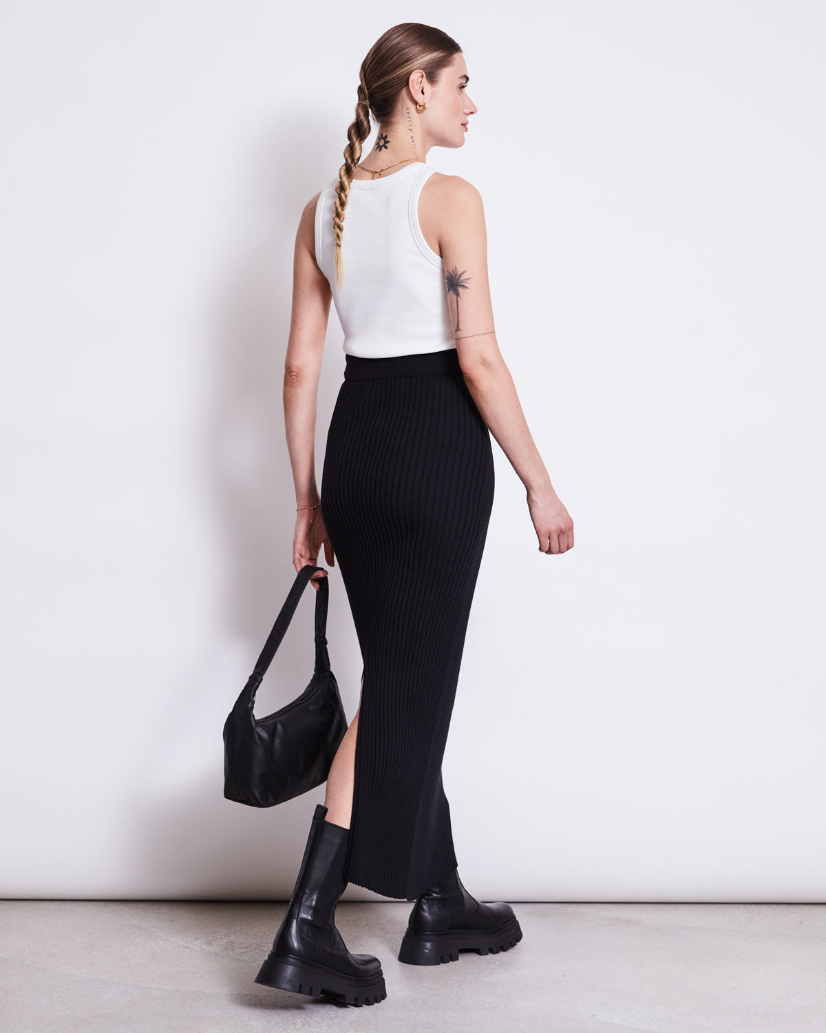 A woman with braided hair wears the jan-n-june MIDI KNIT SKIRT EBBA BLACK GOTS, a white tank top, chunky black boots, and carries a black handbag. She stands against a white background, facing slightly away.