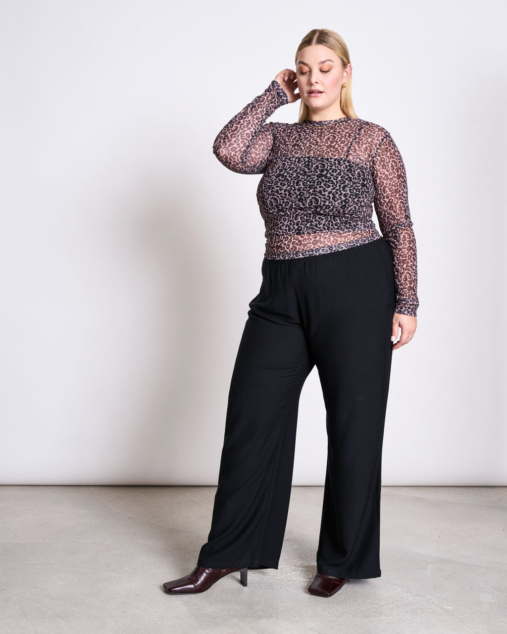 A woman poses indoors against a plain wall, wearing the jan-n-june MESH LONGSLEEVE DORA LEO PRINT top over a black camisole, paired with black wide-leg pants and dark ankle boots. She touches her head and gazes thoughtfully to the side.