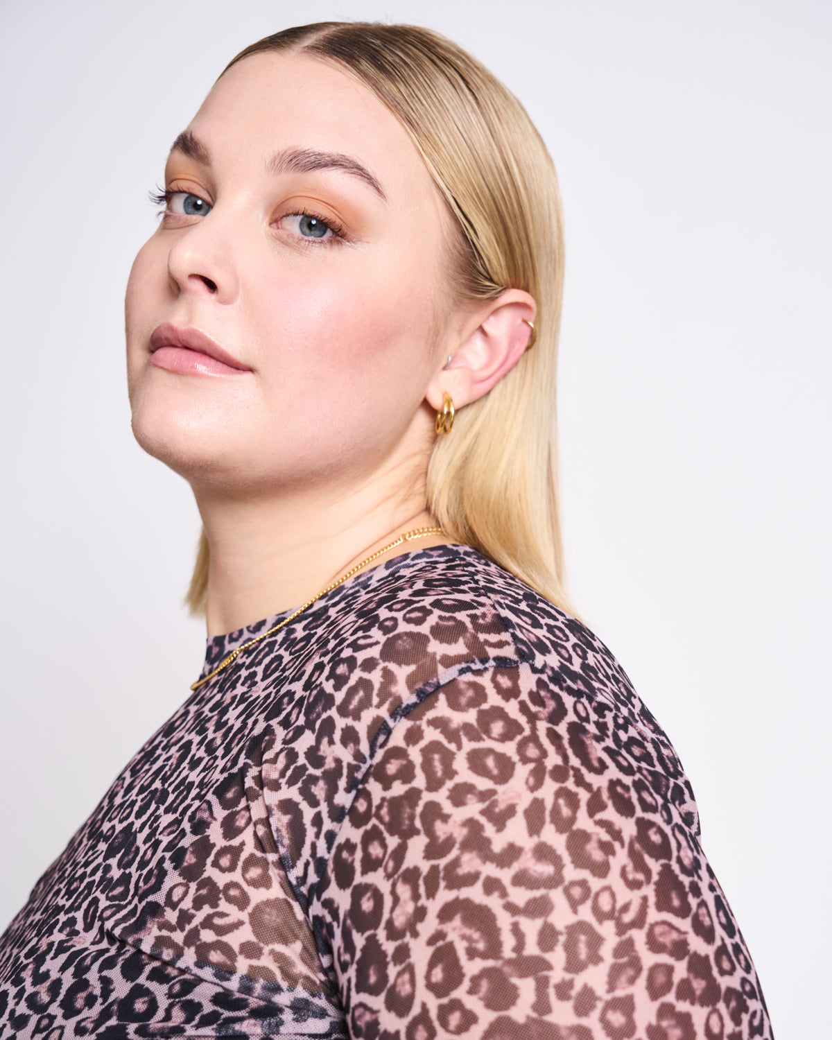 A woman with straight blonde hair and light makeup wears jan-n-junes MESH LONGSLEEVE DORA LEO PRINT, featuring a sheer leopard pattern in recycled polyester, styled with gold hoop earrings against a plain white background.
