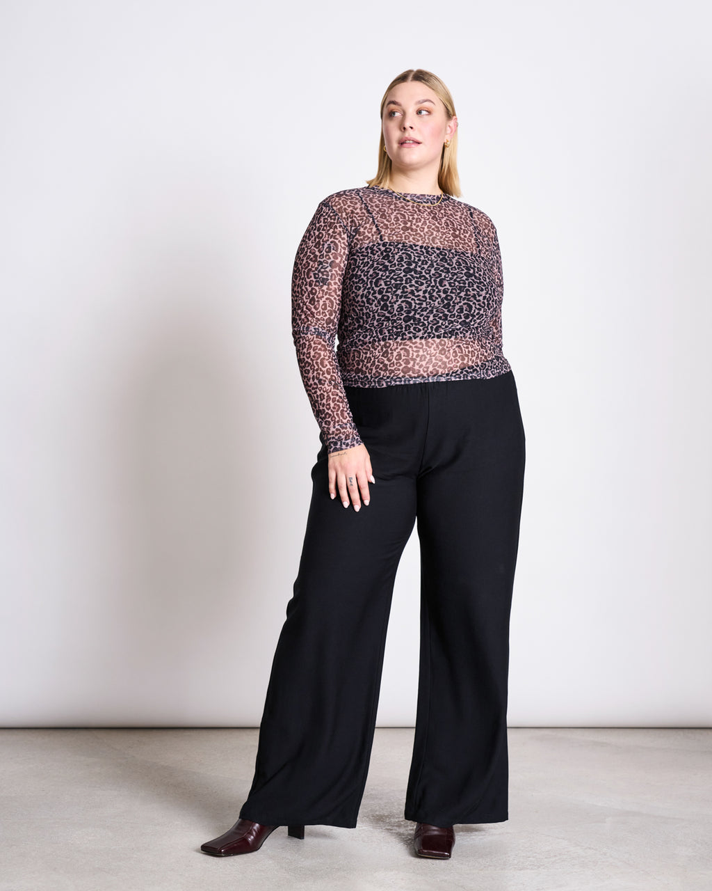 A woman with straight blonde hair poses against a plain background wearing the jan-n-june MESH LONGSLEEVE DORA LEO PRINT top, made from recycled polyester, high-waisted black wide-leg pants, and maroon shoes.