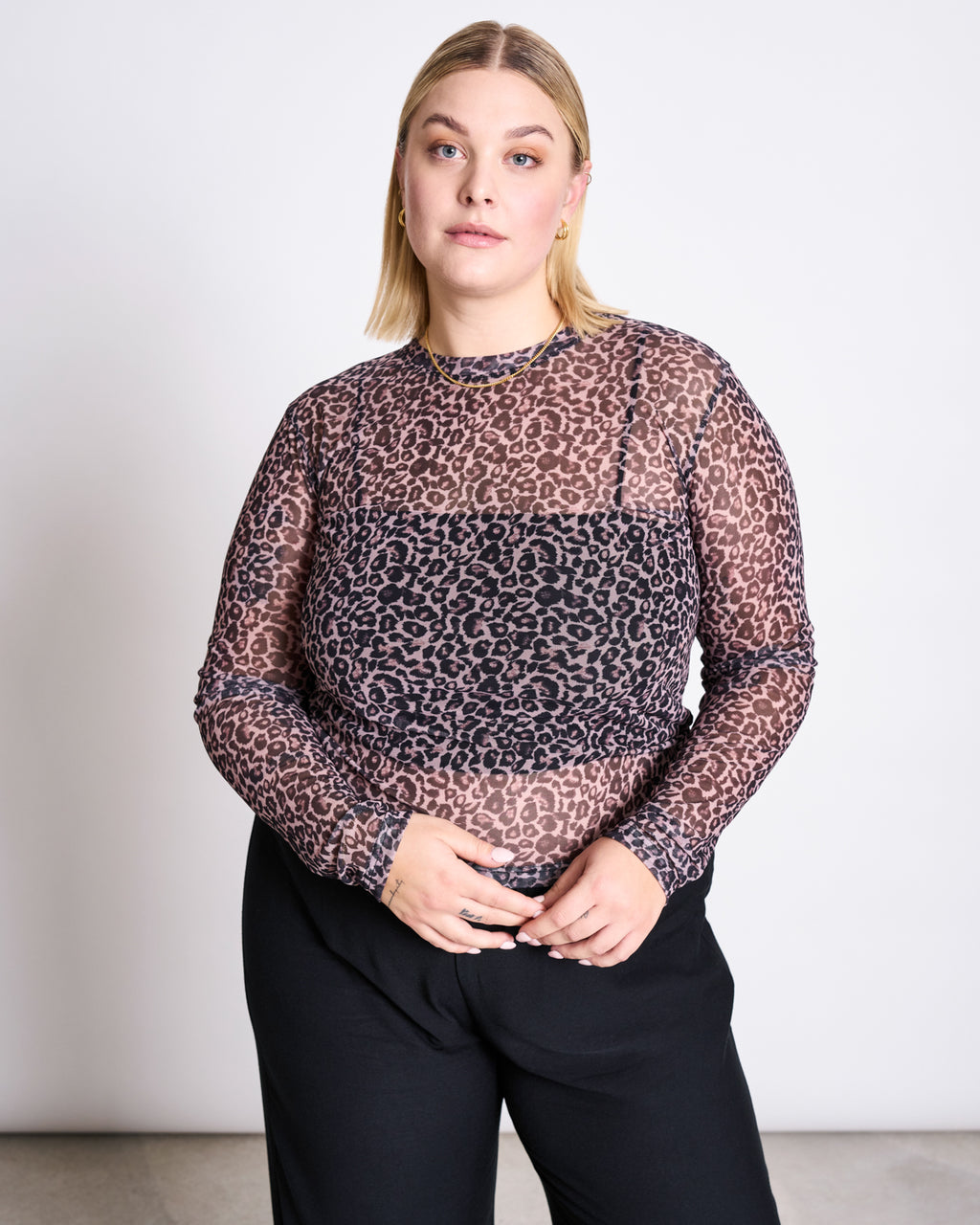 A person with straight blonde hair wears the jan-n-june MESH LONGSLEEVE DORA LEO PRINT over a black undergarment and black pants, standing against a plain light background.
