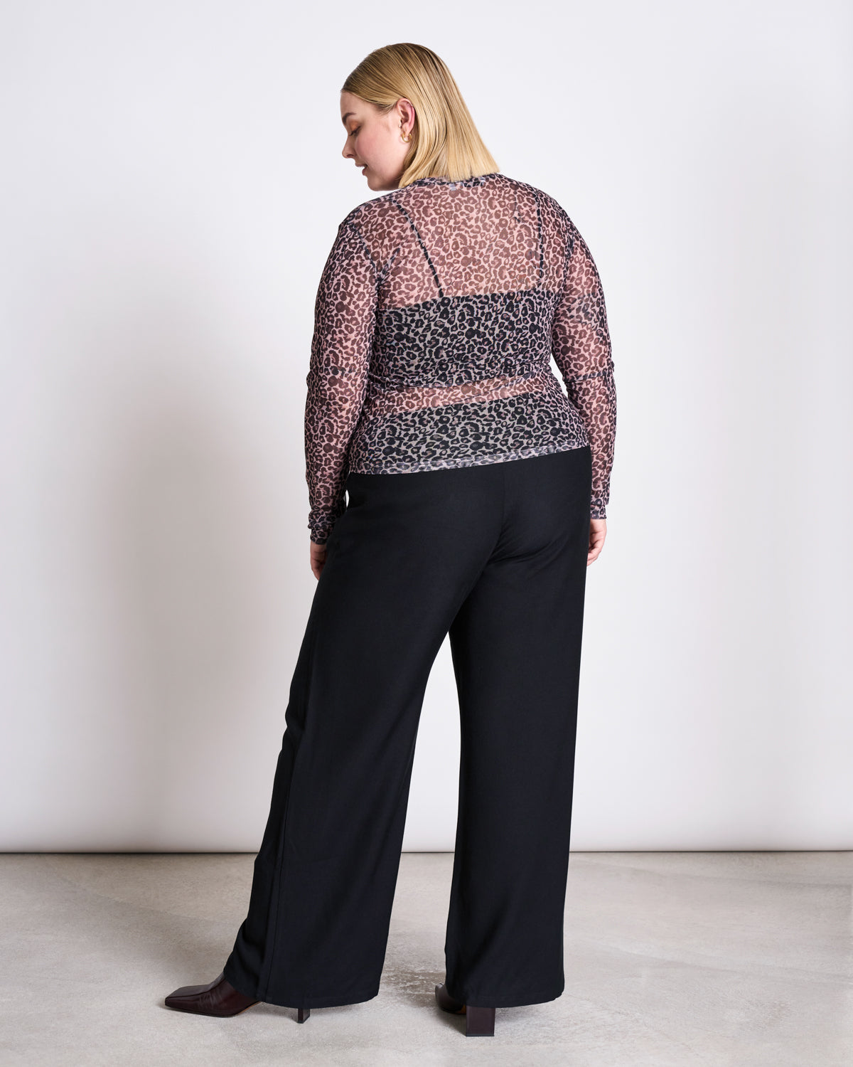 A blonde person stands with their back to the camera in a minimalist space, wearing jan-n-junes MESH LONGSLEEVE DORA LEO PRINT top and loose black pants.