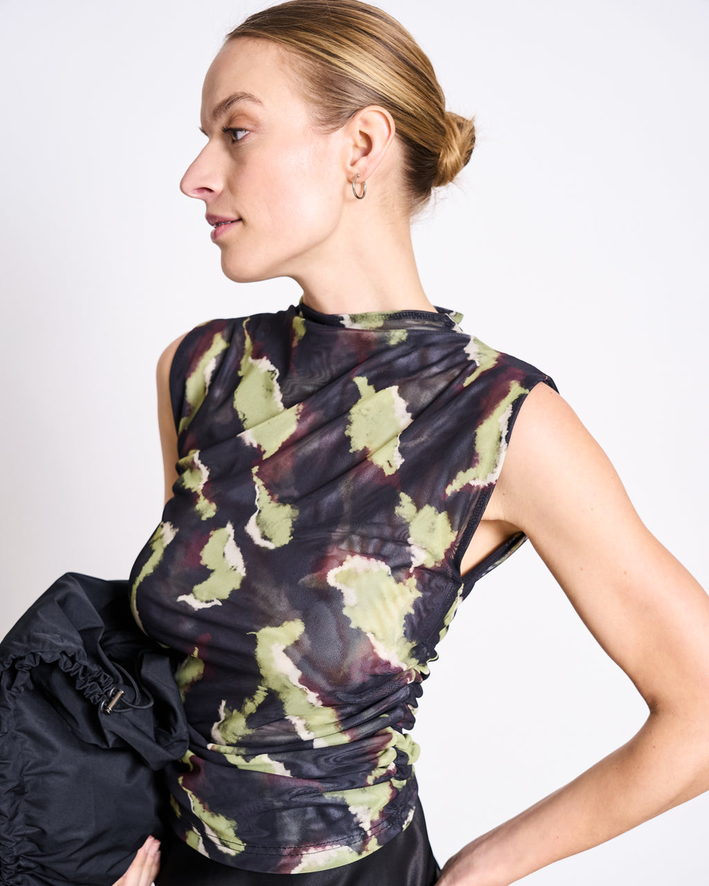 A woman with blonde hair in a bun wears the jan-n-june RUFFLE MESH TOP LAGUNA MOSSFIRE, a sleeveless high-neckline recycled mesh top with a green, black, and maroon abstract pattern. She holds a black ruched bag against a white background.