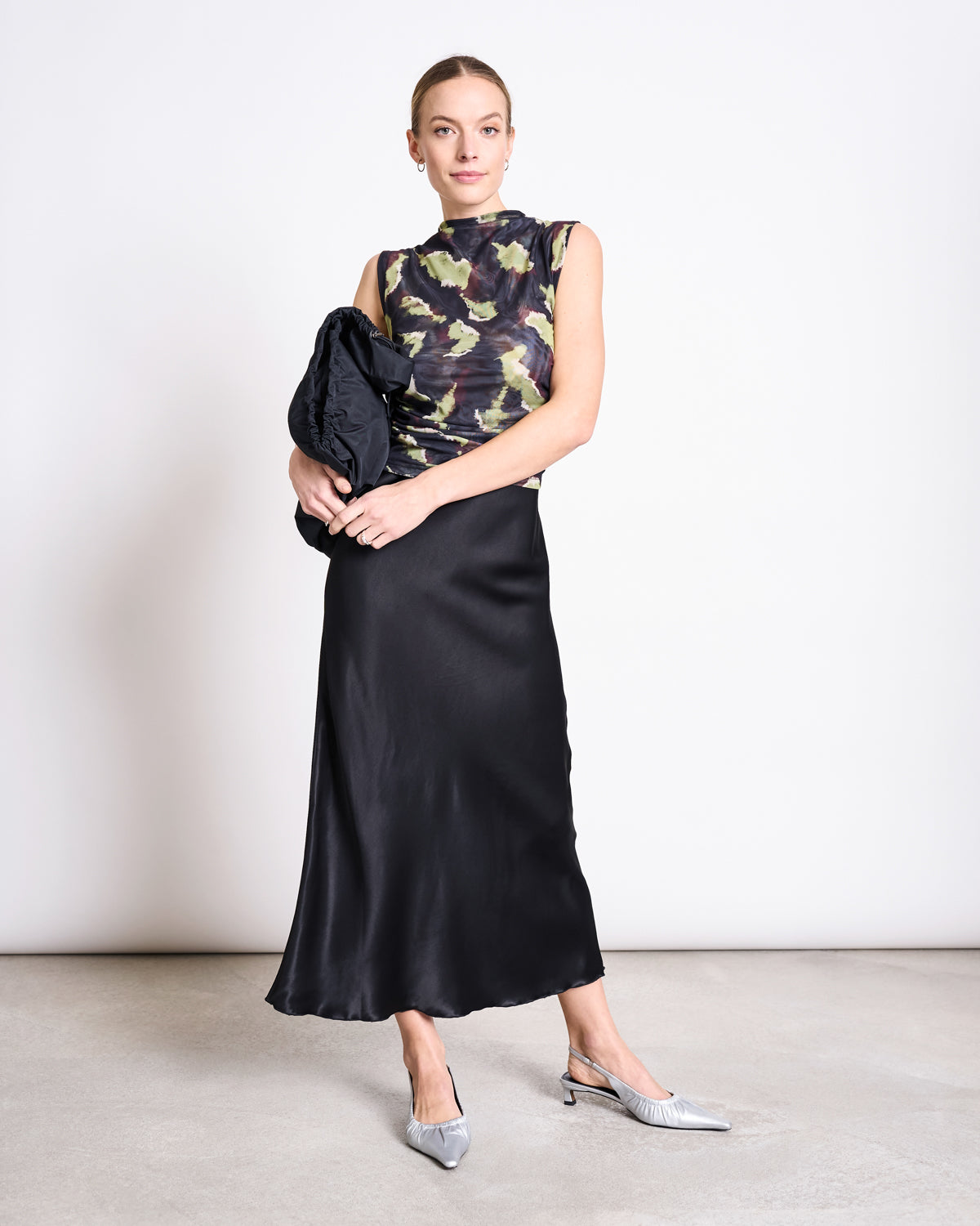 A woman stands against a plain white wall, wearing the jan-n-june RUFFLE MESH TOP LAGUNA MOSSFIRE, a black satin midi skirt, silver pointed heels, and holding a black clutch. She gazes at the camera with a neutral expression.