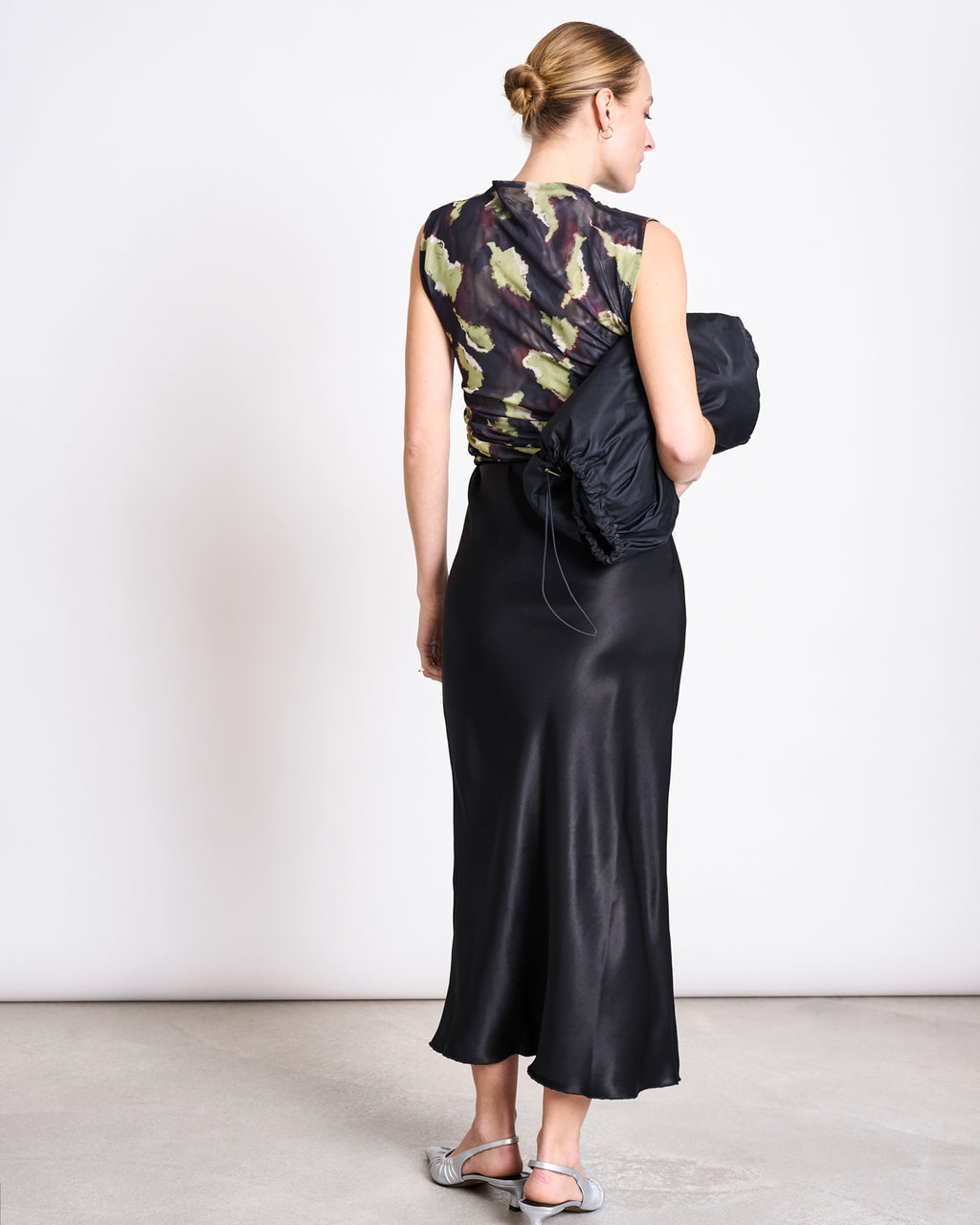 A woman stands facing away, wearing the jan-n-june RUFFLE MESH TOP LAGUNA MOSSFIRE and a long black satin skirt. She holds a black jacket, wears silver slip-on shoes, and has her hair styled in a neat bun.