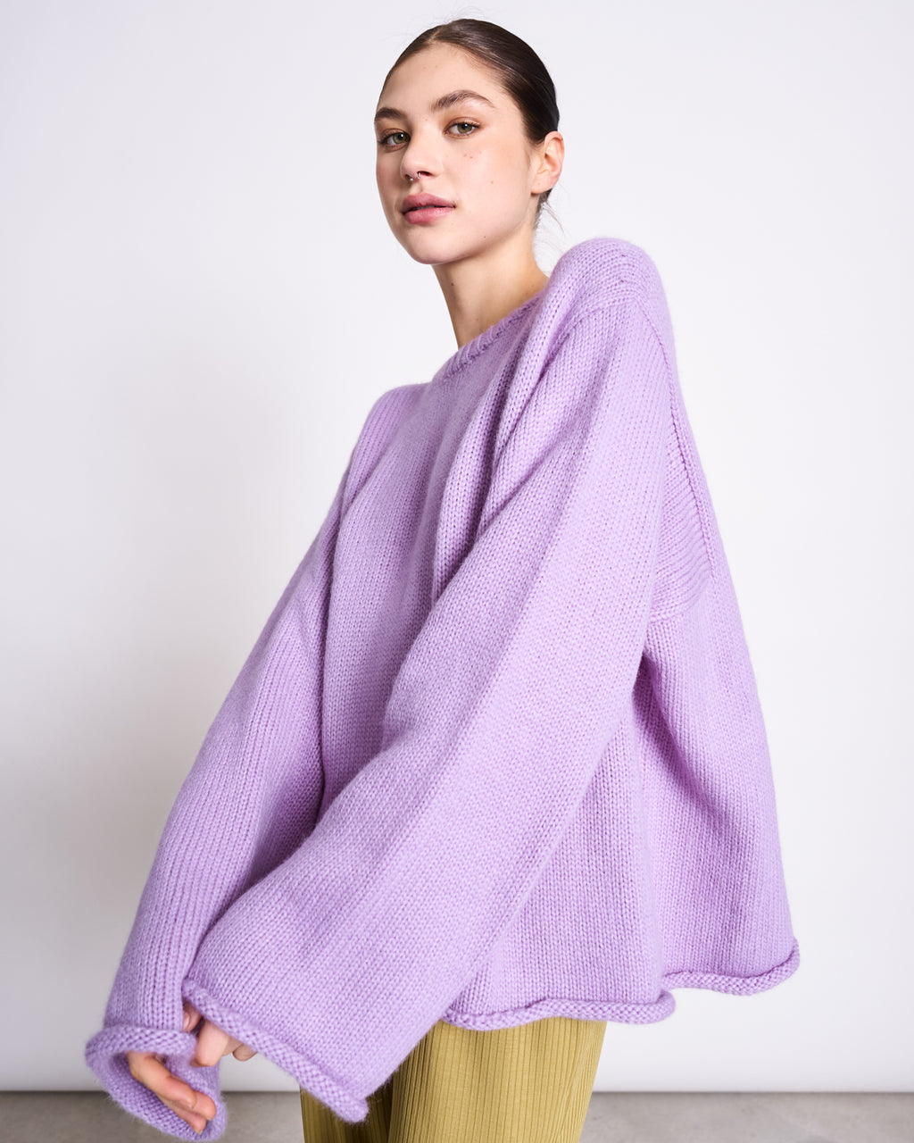 A woman with dark hair pulled back wears the jan-n-june OVERSIZED ALPACA MIX JUMPER WOOLY SONA LAVENDER and yellow pants, standing against a plain white background.