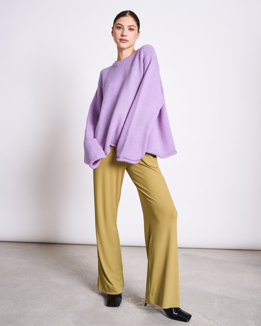 A woman, hair pulled back, confidently poses against a plain background in the jan-n-june OVERSIZED ALPACA MIX JUMPER WOOLY SONA LAVENDER, paired with mustard-yellow wide-leg pants and black shoes.