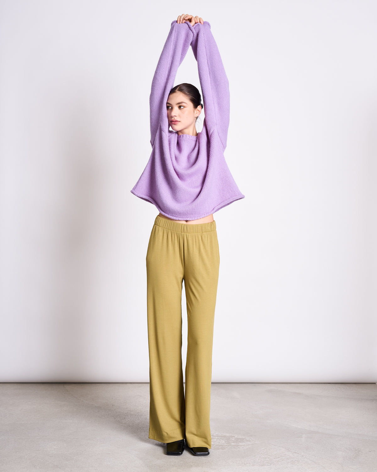 A woman stands indoors on a concrete floor against a plain white background, wearing the jan-n-june OVERSIZED ALPACA MIX JUMPER WOOLY SONA LAVENDER with wide-leg mustard yellow pants and black shoes, arms raised overhead.