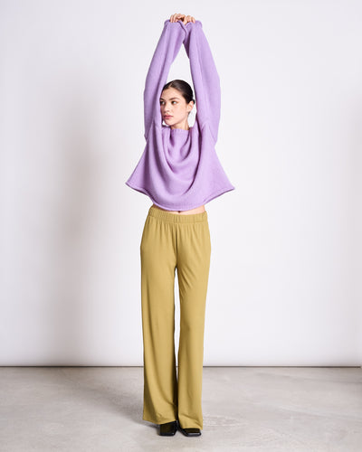 A woman stands indoors on a concrete floor against a plain white background, wearing the jan-n-june OVERSIZED ALPACA MIX JUMPER WOOLY SONA LAVENDER with wide-leg mustard yellow pants and black shoes, arms raised overhead.