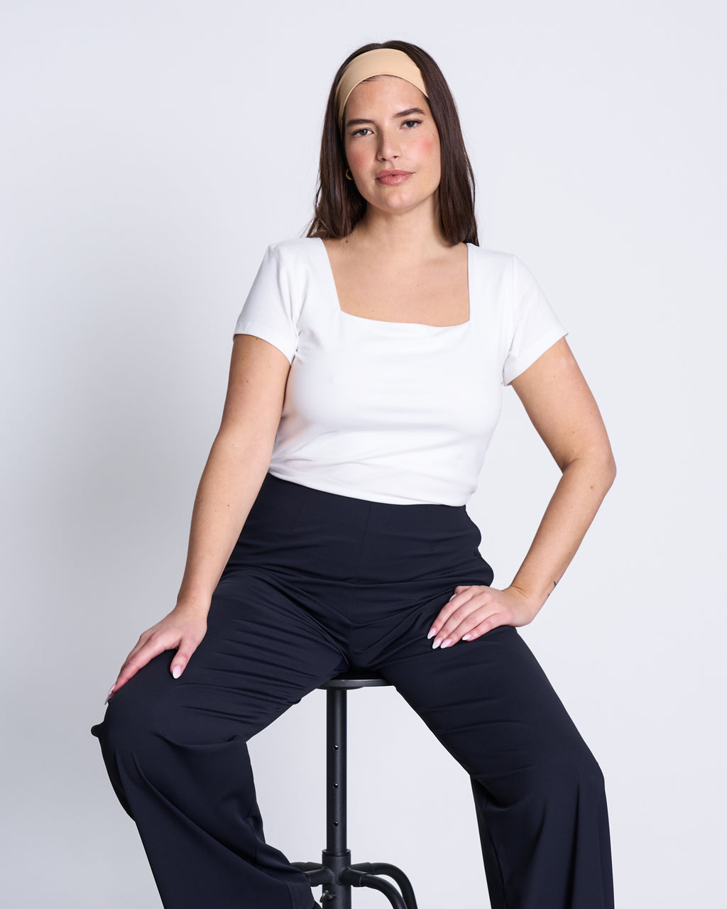 A woman with long brown hair wears the jan-n-june T-SHIRT ARIMA WHITE GOTS, a super soft square neck short-sleeve top, paired with black pants and a tan headband as she sits confidently on a stool against a plain white background.