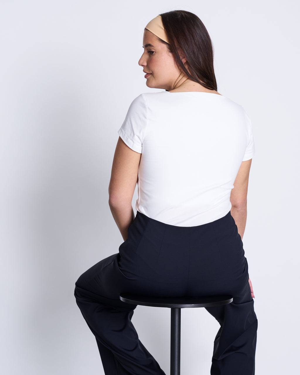 A woman with long dark hair, dressed in the jan-n-june T-SHIRT ARIMA WHITE GOTS and black pants, sits on a stool facing away from the camera against a plain light background.