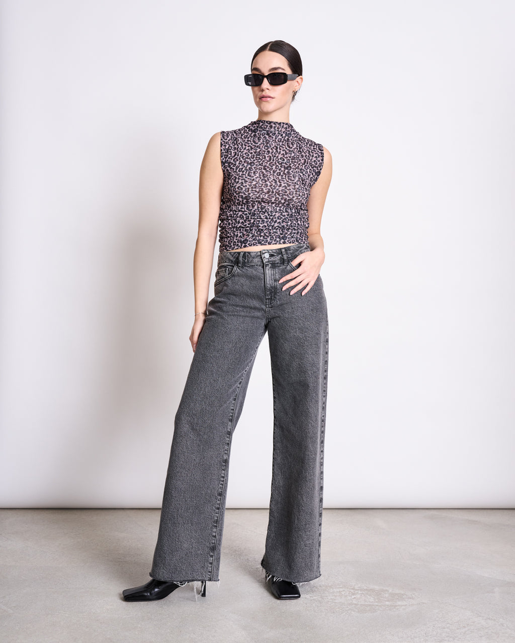 A woman in jan-n-june’s RUFFLE TOP LAGUNA LEO PRINT, black sunglasses, high-waisted wide-leg gray jeans, and black heeled boots stands on a concrete floor against a plain white background.