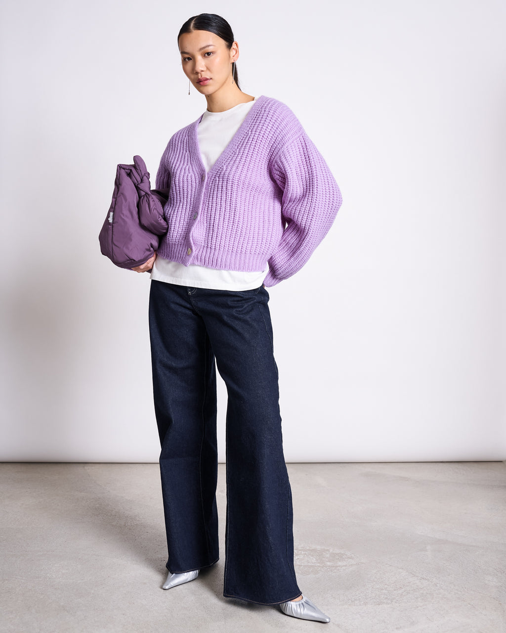 A woman poses against a plain background in the jan-n-june ALPACA MIX CARDIGAN WOOLY LENA LAVENDER, paired with a white top, dark wide-leg jeans, shiny silver pointed shoes, and a matching purple handbag for timeless style.