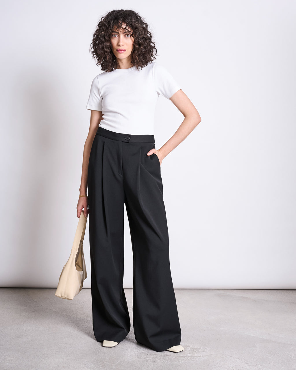 A person with curly hair wears jan-n-junes WIDE PANTS GIZA BLACK, a white short-sleeve top, beige handbag, and light shoes, standing against a plain background. The high-waisted pants are made from recycled polyester.
