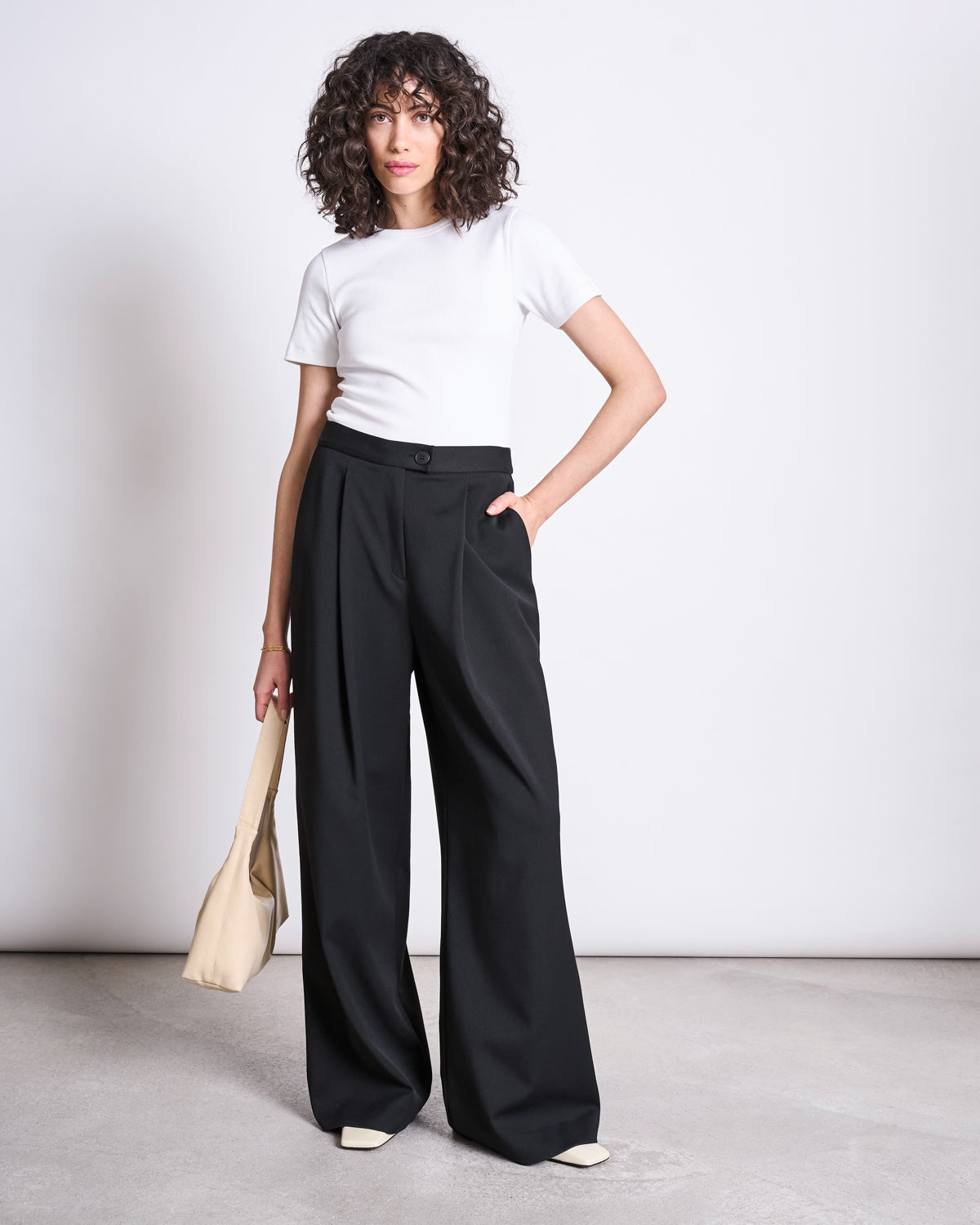 A person with curly hair wears jan-n-junes WIDE PANTS GIZA BLACK, a white short-sleeve top, beige handbag, and light shoes, standing against a plain background. The high-waisted pants are made from recycled polyester.