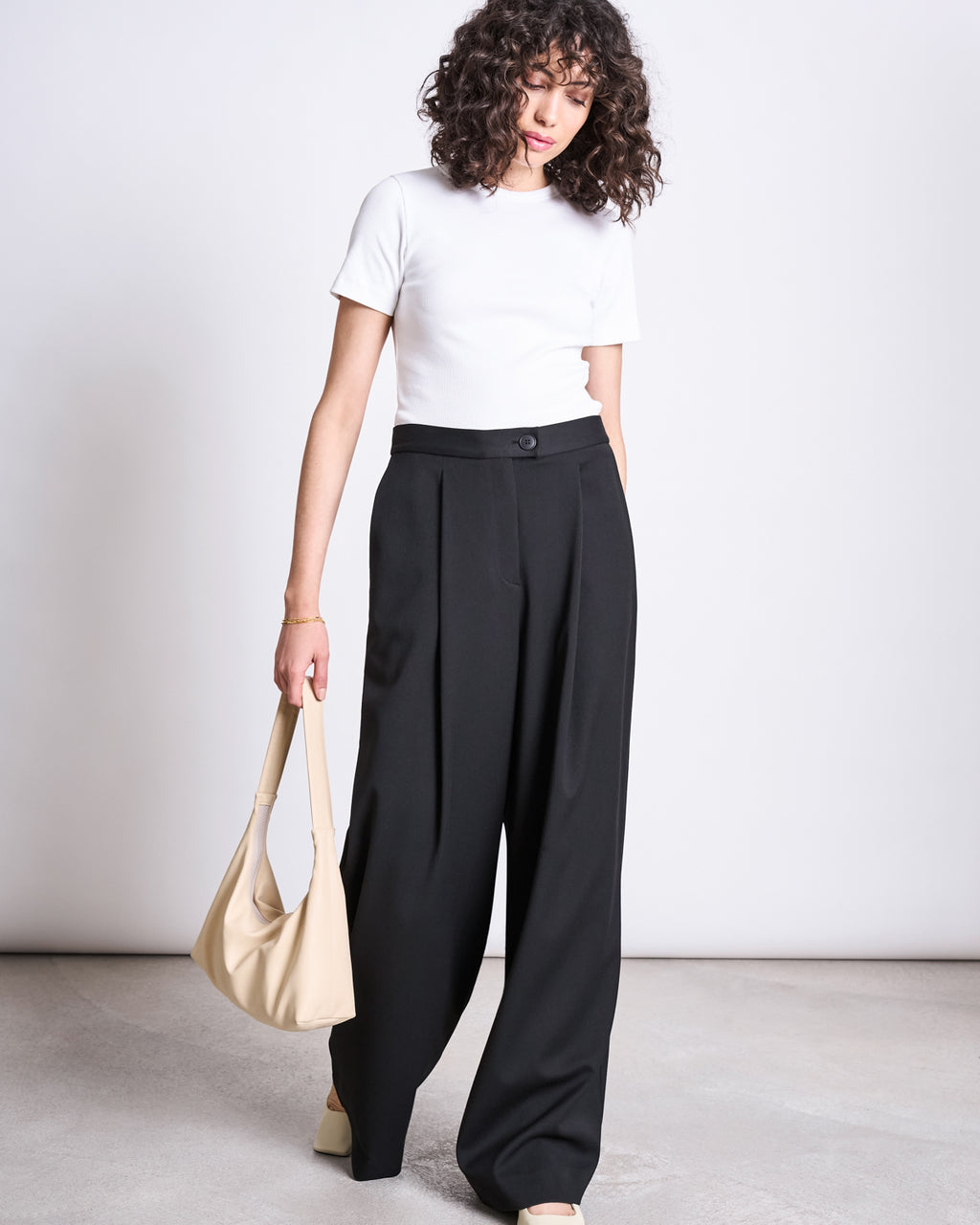 A woman with curly hair wears jan-n-june’s WIDE PANTS GIZA BLACK with a white short-sleeve top and cream shoes, holding a beige handbag against a plain white background.