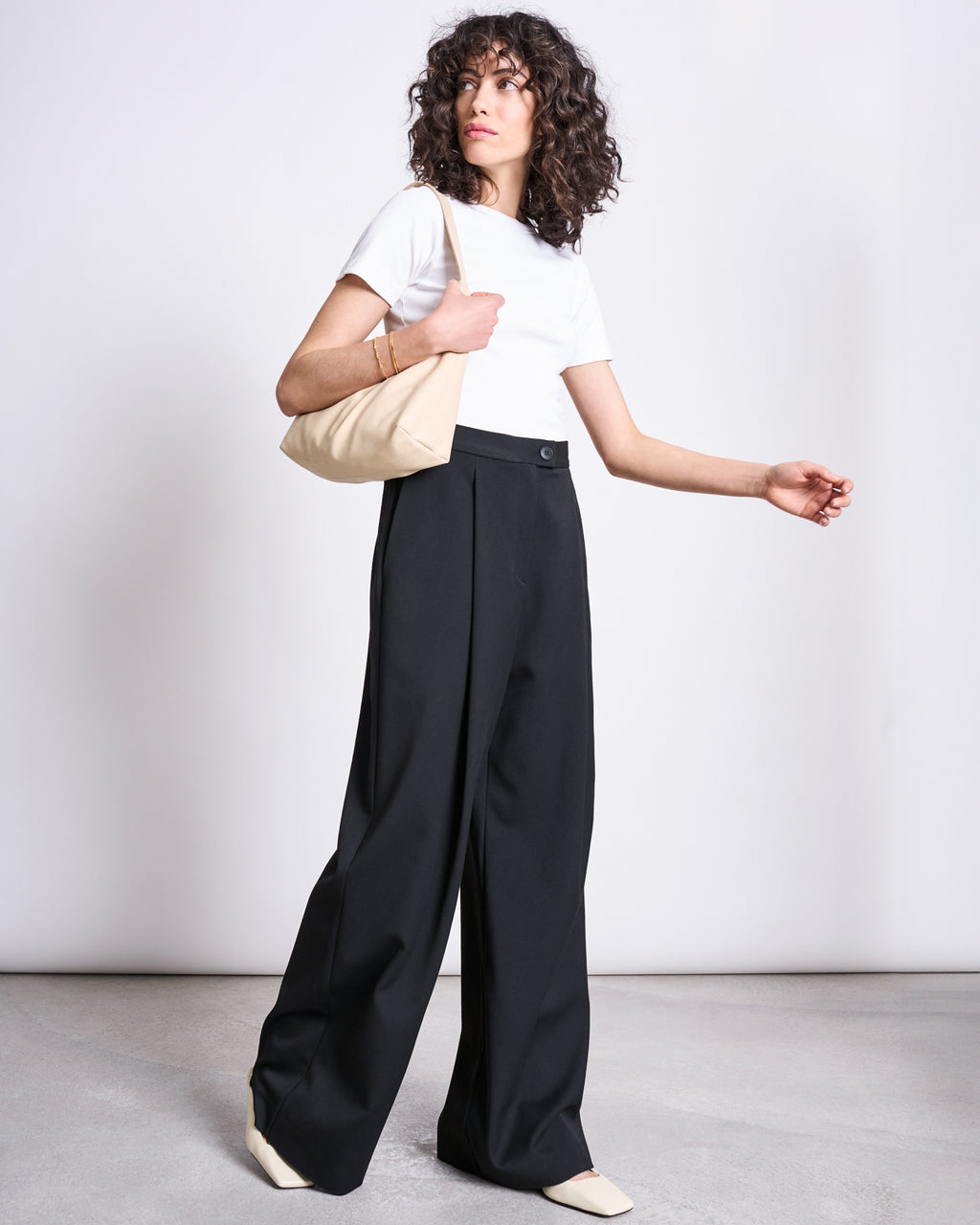 A person with curly hair stands against a plain background, wearing jan-n-june’s WIDE PANTS GIZA BLACK, a white t-shirt, cream shoes, and holding a beige shoulder bag.