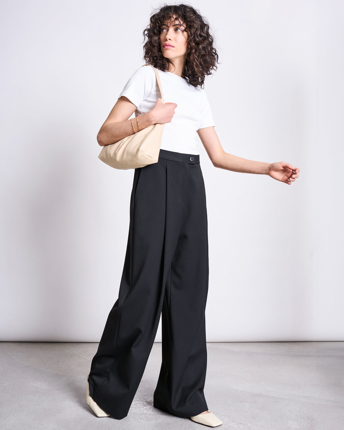 A person with curly hair stands against a plain background, wearing jan-n-june’s WIDE PANTS GIZA BLACK, a white t-shirt, cream shoes, and holding a beige shoulder bag.
