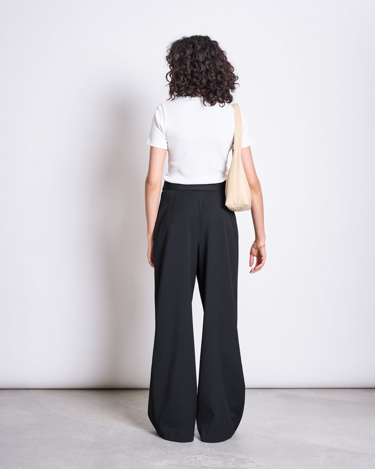 A person with curly hair faces away, wearing jan-n-junes WIDE PANTS GIZA BLACK—high-waisted, wide-leg pants made from recycled polyester—with a white short-sleeve top and a beige shoulder bag against a plain light background.