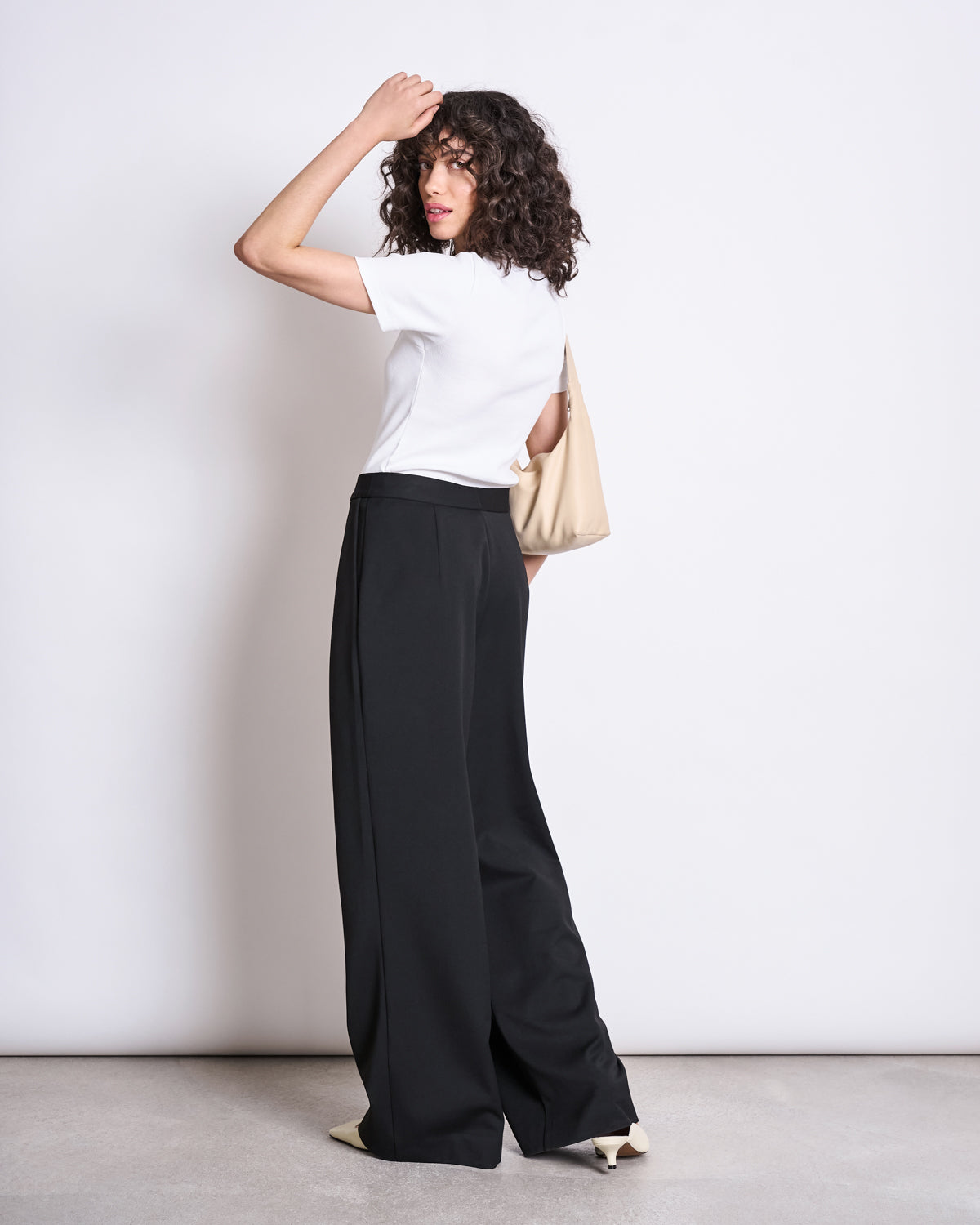 A woman with curly hair wears jan-n-junes WIDE PANTS GIZA BLACK, a white tee, white heels, and carries a beige bag. She stands facing away, looking over her shoulder against a plain white background.