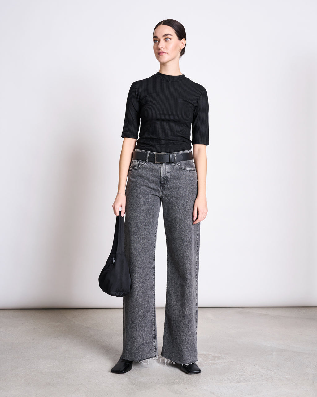 A woman stands against a plain background wearing the jan-n-june LYOCELL RIB T-SHIRT NINA BLACK, high-waisted gray wide-leg jeans, a black belt, pointed shoes, and holding a black bag by her side.