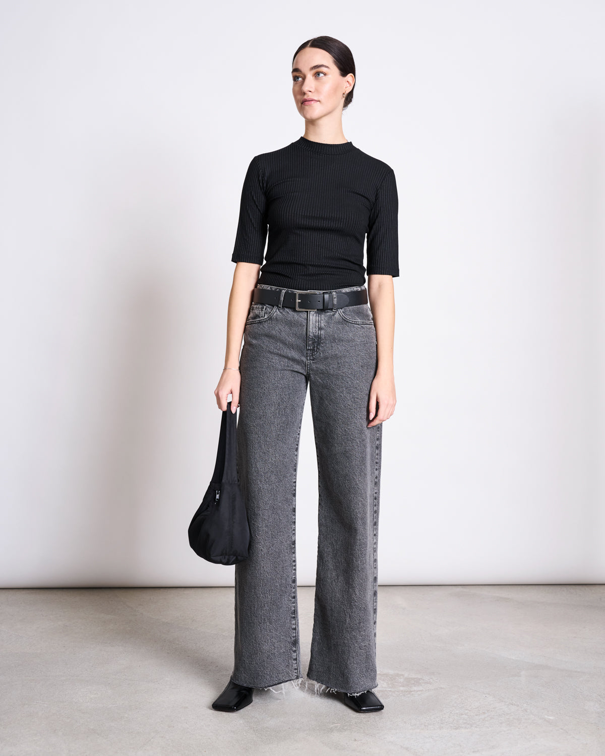 A woman stands against a plain background wearing the jan-n-june LYOCELL RIB T-SHIRT NINA BLACK, high-waisted gray wide-leg jeans, a black belt, pointed shoes, and holding a black bag by her side.