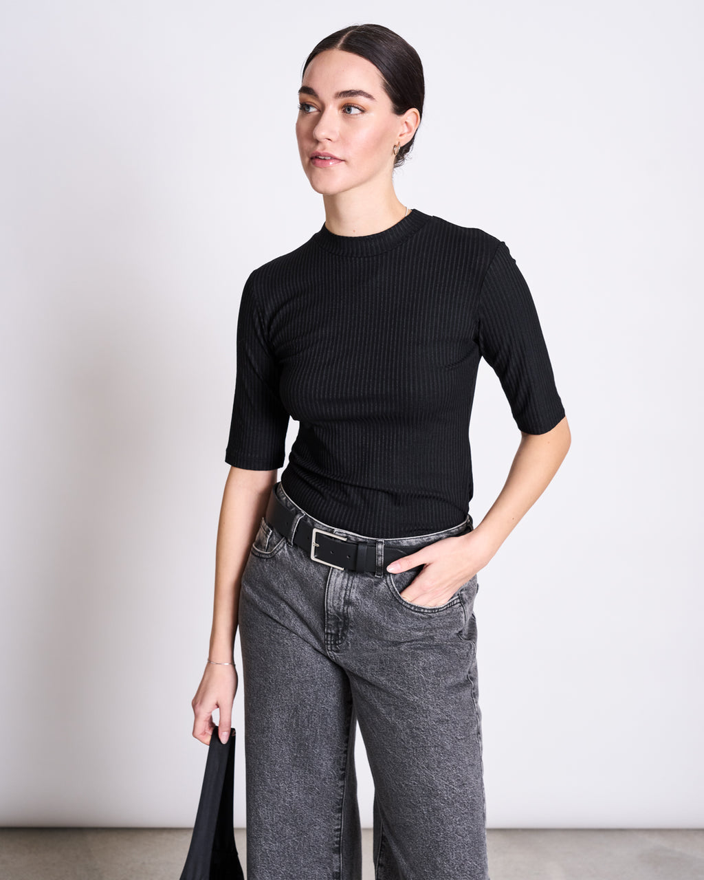 A woman with dark hair pulled back wears the jan-n-june LYOCELL RIB T-SHIRT NINA BLACK, gray high-waisted jeans, and a black belt. She stands against a white background, holding a black bag and looking to the side.