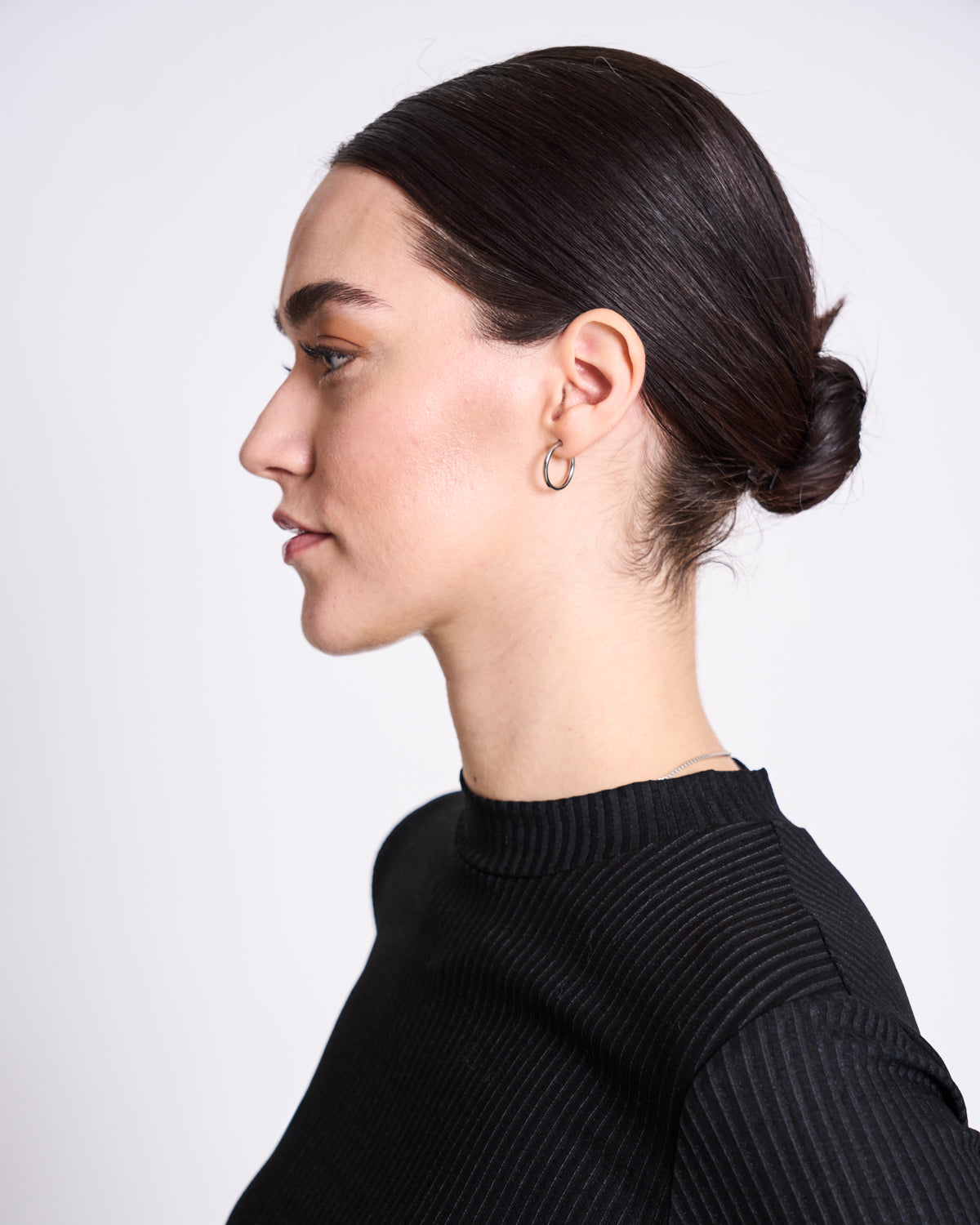 A woman with dark hair in a low bun, wearing the jan-n-june LYOCELL RIB T-SHIRT NINA BLACK and small hoop earrings, is shown in profile against a plain white background.