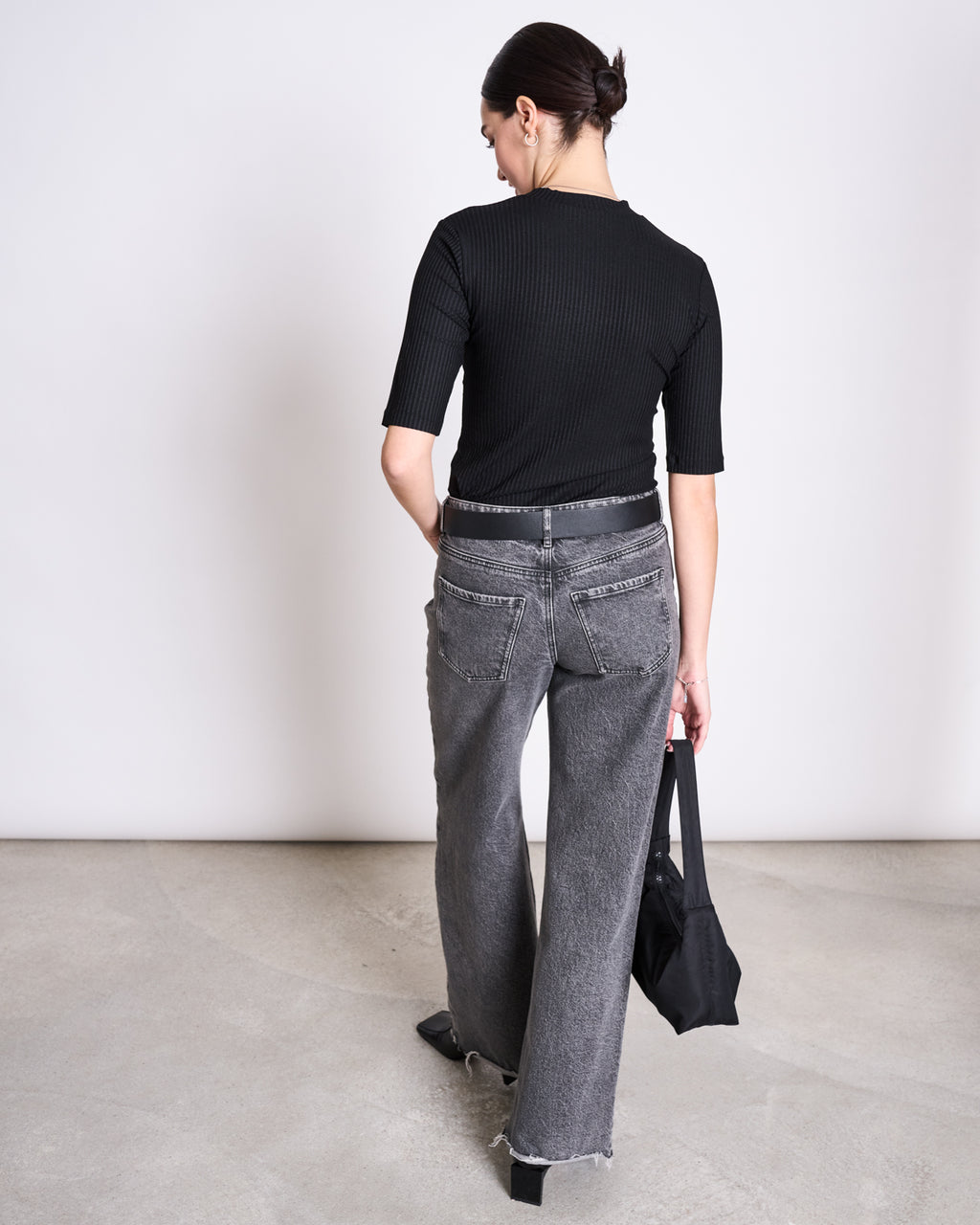 A woman with dark hair in a low bun stands indoors on a concrete floor, wearing the jan-n-june LYOCELL RIB T-SHIRT NINA BLACK and gray wide-leg jeans with frayed hems, holding a black handbag in her left hand.
