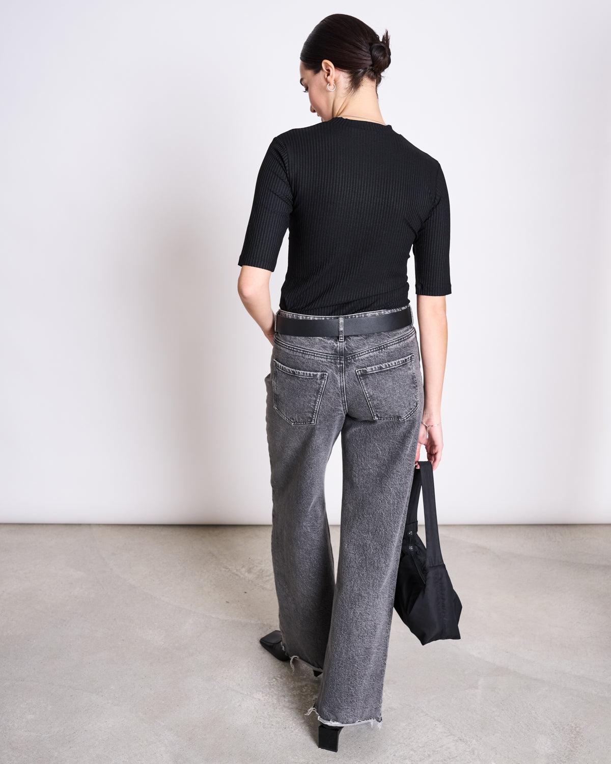 A woman with dark hair in a low bun stands indoors on a concrete floor, wearing the jan-n-june LYOCELL RIB T-SHIRT NINA BLACK and gray wide-leg jeans with frayed hems, holding a black handbag in her left hand.