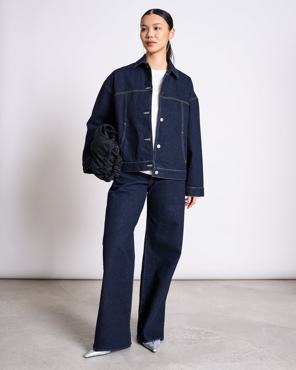 A woman stands against a plain background wearing the jan-n-june RELAXED DENIM JACKET VILME DARK BLUE DENIM GOTS, styled with matching wide-leg jeans, a white top, silver pointed-toe shoes, and a black scrunched handbag.