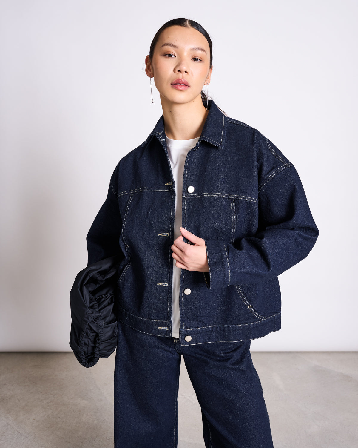 A woman stands indoors against a plain backdrop, wearing the jan-n-june RELAXED DENIM JACKET VILME DARK BLUE DENIM GOTS and matching wide-leg jeans over a white shirt, holding a black item in her left hand as she gazes confidently at the camera.