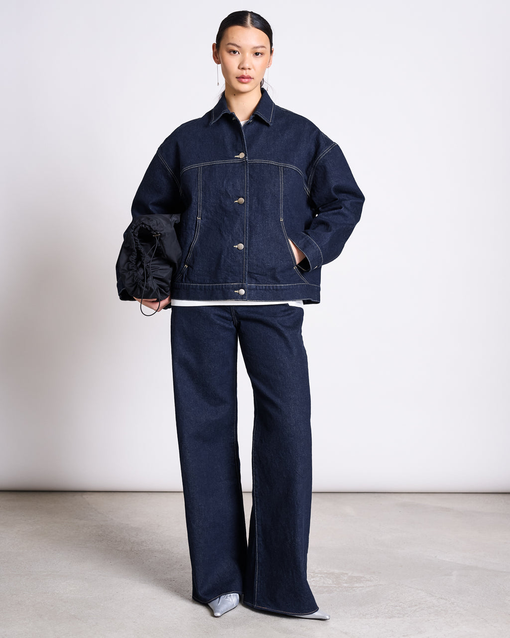 A person stands against a plain background, wearing the jan-n-june RELAXED DENIM JACKET VILME DARK BLUE DENIM GOTS with matching wide-leg jeans, holding a black clutch and wearing pointed silver shoes.